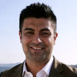 A man with short black hair, wearing a brown suit jacket and white shirt, smiles outdoors with a clear blue sky and distant hills in the background.